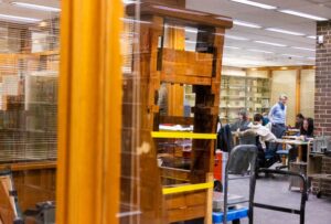 A printing press is shown in the foreground as a professor leads a class of students in the background.