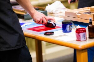 A student's hand is shown spreading red ink onto a roller for a print on a letterpress.